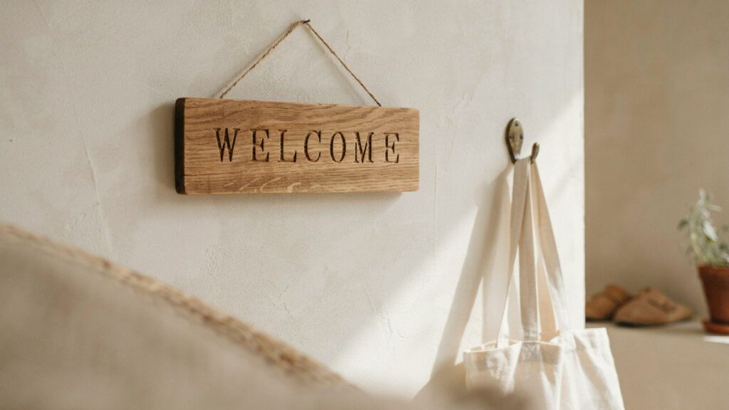 An entryway with a hand-carved oak welcome sign, cream plaster walls, warm morning daylight, and a linen tote on a brass hook